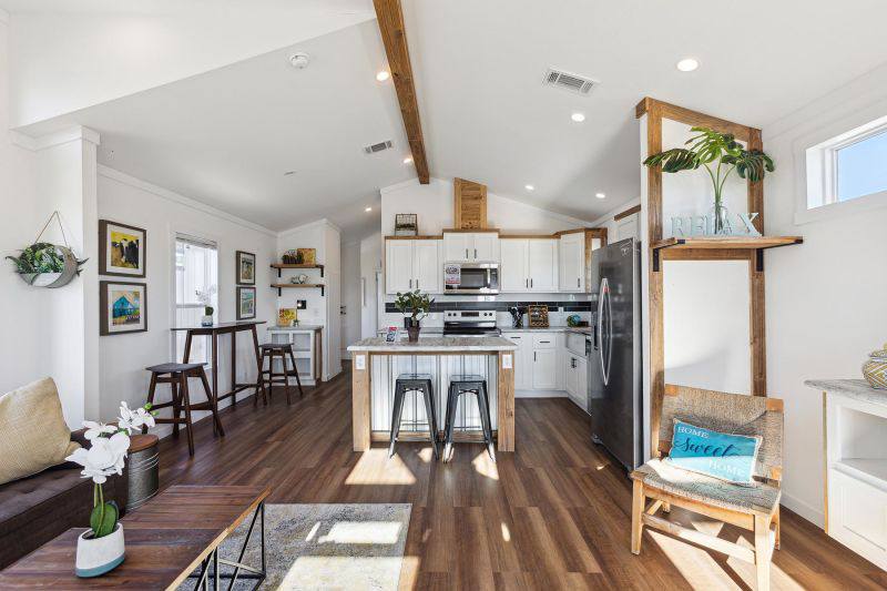 Modern interior of a spacious tiny home featuring a bright white kitchen with a central island, white cabinetry, and a sleek black backsplash, flowing into a cozy living area with rich wood flooring, exposed wooden beams on the vaulted ceiling, and comfortable seating, creating an inviting and stylish living space.