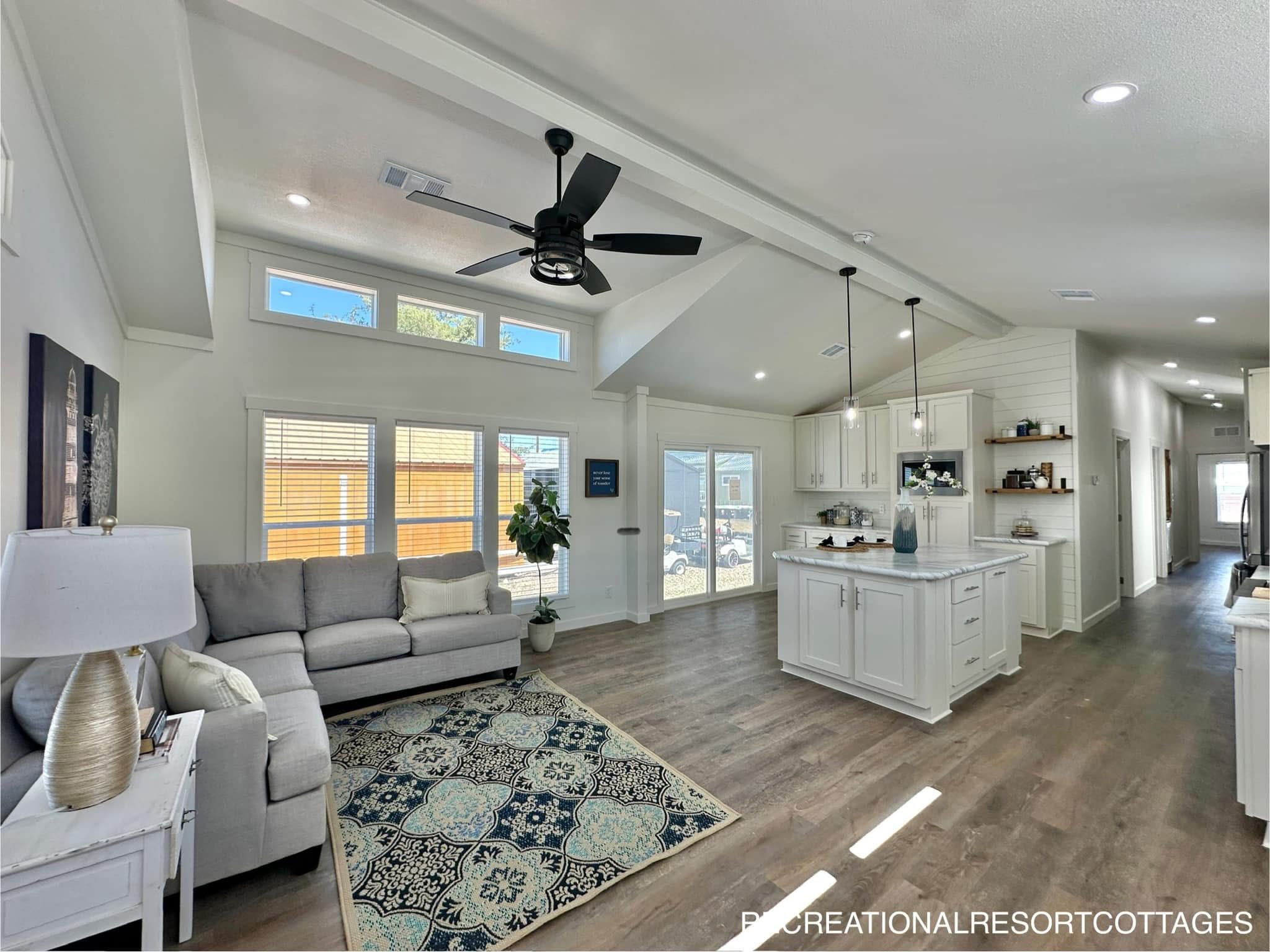 Spacious, modern living room and kitchen with white walls and cabinetry. A gray sofa, patterned rug, ceiling fan, and large windows create an airy feel.