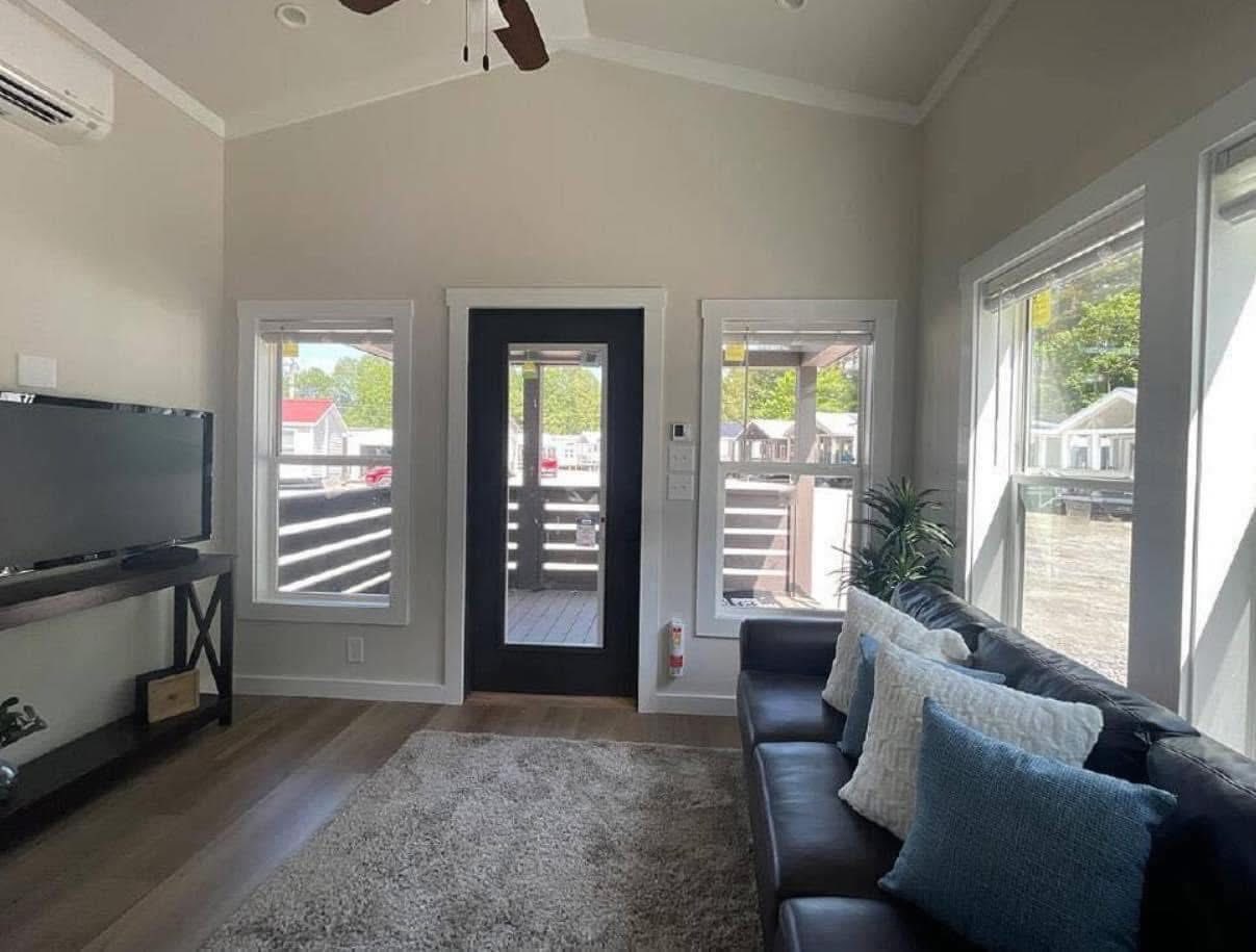 Bright and airy living area of a modern tiny home, featuring a dark leather sofa with textured white and blue throw pillows, a large gray area rug, a black entertainment console with a flat-screen TV, and a vaulted ceiling with a ceiling fan, all illuminated by natural light streaming through multiple large windows and a glass-paneled front door that opens to a covered porch, with a glimpse of other tiny homes in the background.