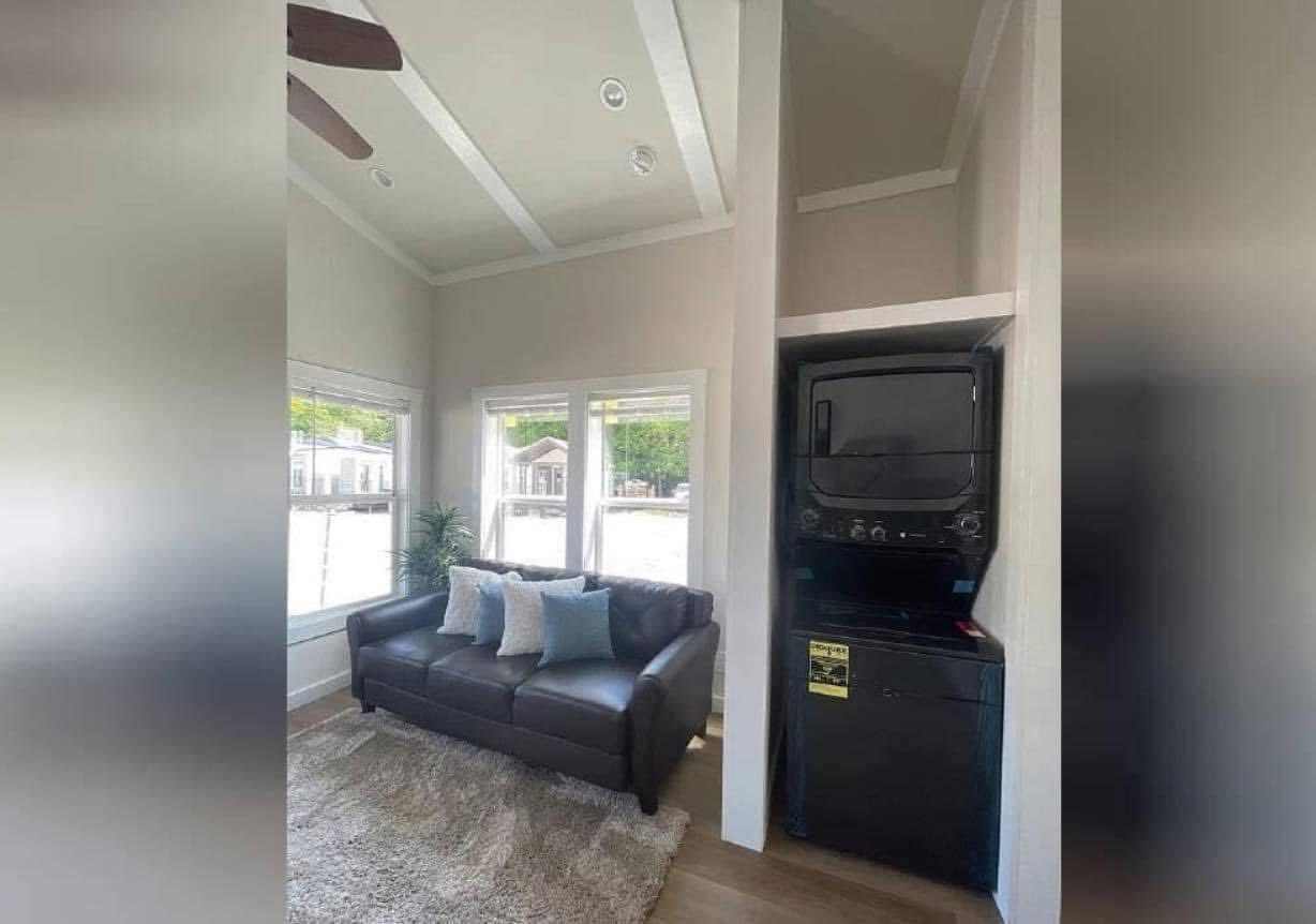 Cozy living room in a compact tiny home featuring a dark brown leather sofa with light blue and white accent pillows, a plush beige area rug, and a stacked black washer and dryer unit in a built-in closet, all under a vaulted ceiling with a ceiling fan and recessed lighting, with large windows offering views of the outdoors.
