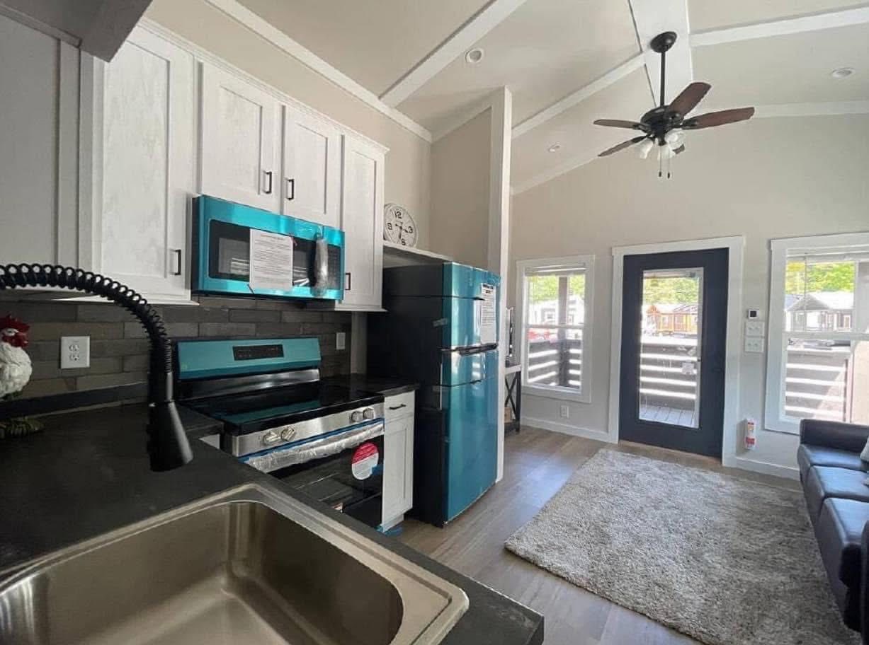 Modern tiny home interior showcasing a vibrant turquoise kitchen with white cabinets, a stainless steel sink, a black range, and a matching turquoise refrigerator, all under a vaulted ceiling with a ceiling fan, adjacent to a living area with a dark sofa, a plush gray rug, and large windows offering views of other tiny homes.