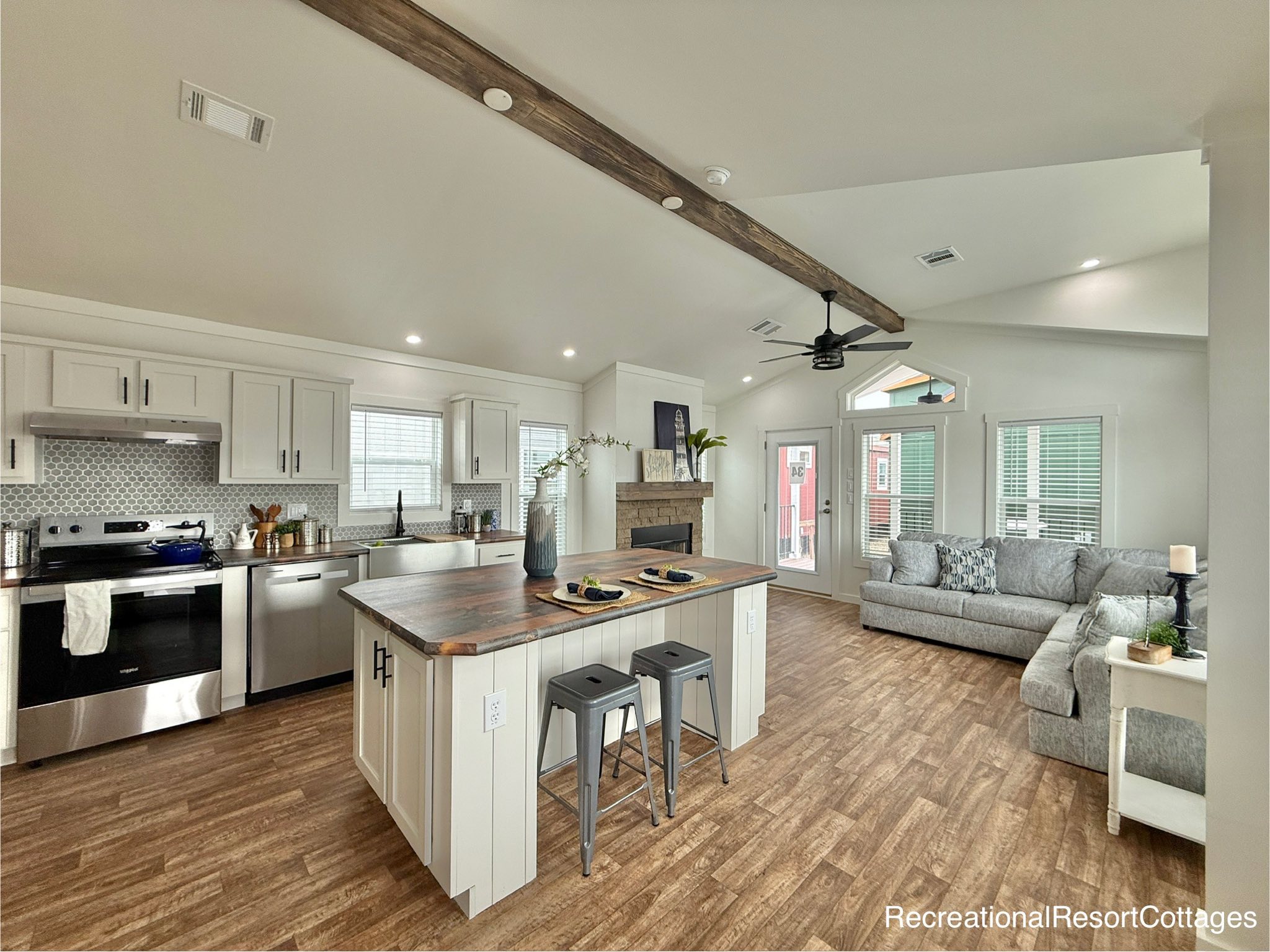 Spacious open-concept living and kitchen area in a modern modular home featuring a grey upholstered sectional sofa with patterned pillows, large windows, a white kitchen island with a distressed wood countertop, white shaker cabinets, stainless steel appliances, a dark farmhouse sink, and a rustic wooden beam accent on the vaulted ceiling with a black ceiling fan, all on warm-toned wood-look laminate flooring.