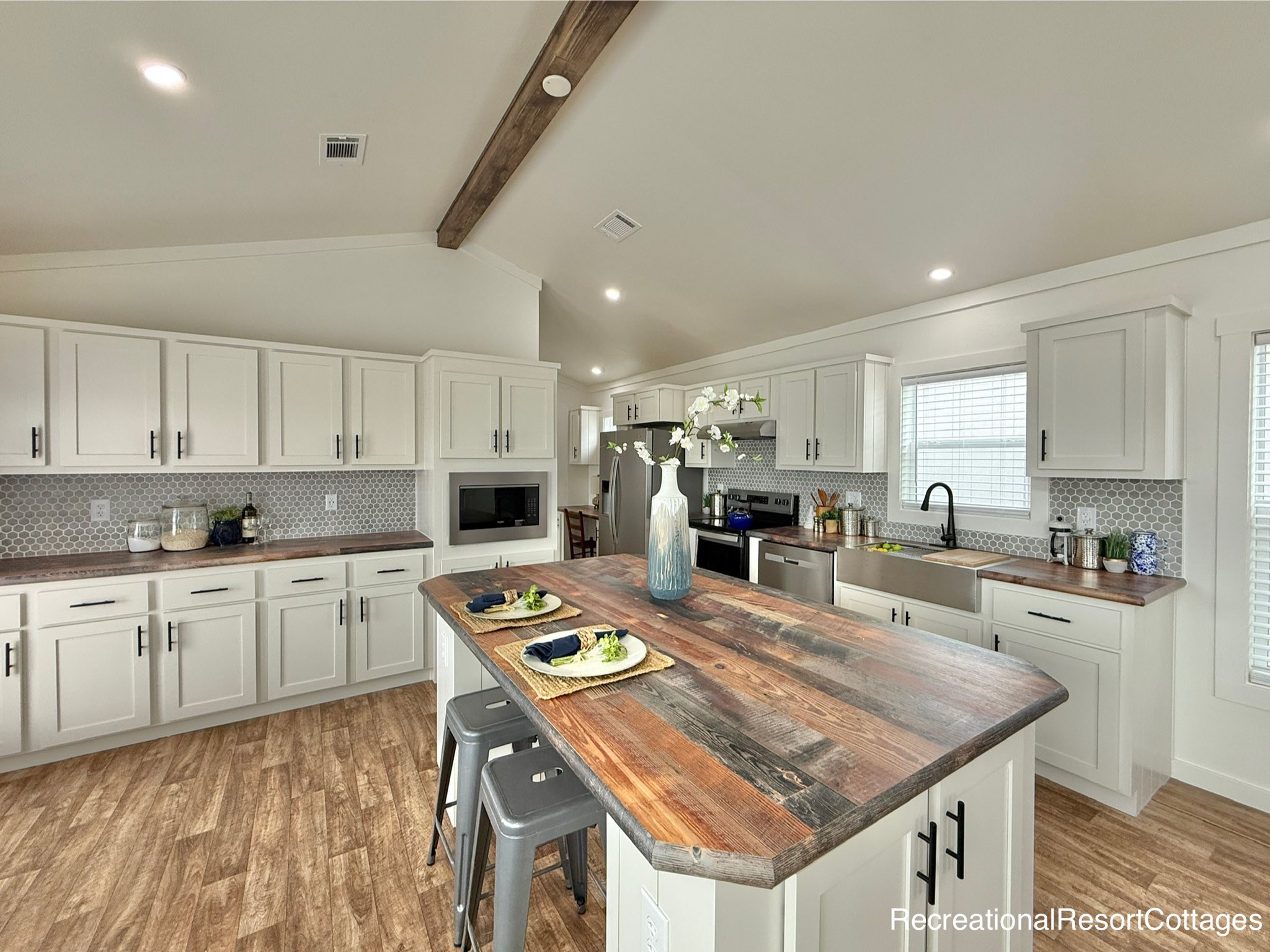 Stunning open-concept modular home kitchen featuring a large, distressed wood island with seating, white shaker cabinets, a grey hexagon tile backsplash, stainless steel appliances including a built-in microwave and a double-door refrigerator, a farmhouse sink, and a rustic wooden beam accent on the vaulted ceiling, all complemented by warm wood-look laminate flooring.