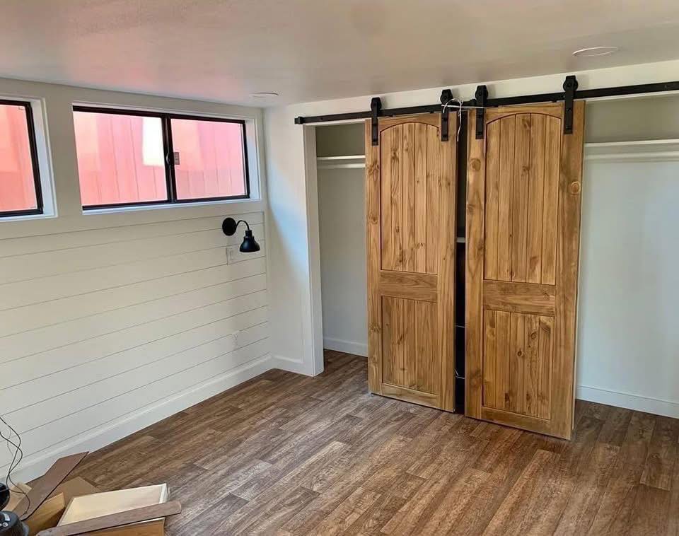 Bright room with wood sliding barn doors on a closet. White shiplap walls, laminate wood floors, and a modern black wall lamp under narrow windows.
