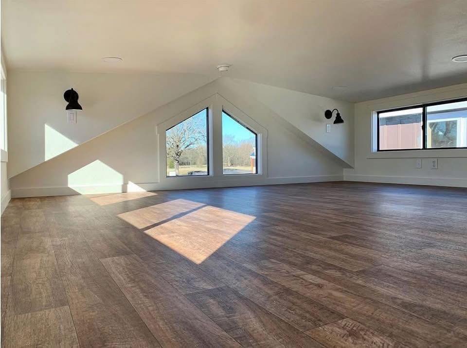 A bright attic space with wooden flooring, a triangular window, and two wall lamps. Sunlight streams in, creating a warm, peaceful ambiance.