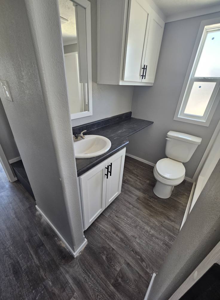 Small bathroom with a modern design. It features a white sink with dark countertop, white cabinets, a toilet, and a window, all on wood-look flooring.
