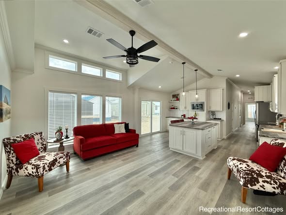Spacious open-concept tiny home interior with high vaulted ceilings, modern ceiling fan, recessed lighting, large windows with blinds flooding natural light, bright white shaker kitchen cabinets, large center island with quartz countertop, stainless steel appliances, cozy red sofa, cow print accent chairs, gray wood-look flooring, hallway leading to bedrooms – luxurious recreational resort cottage design for tiny house and vacation home inspiration.