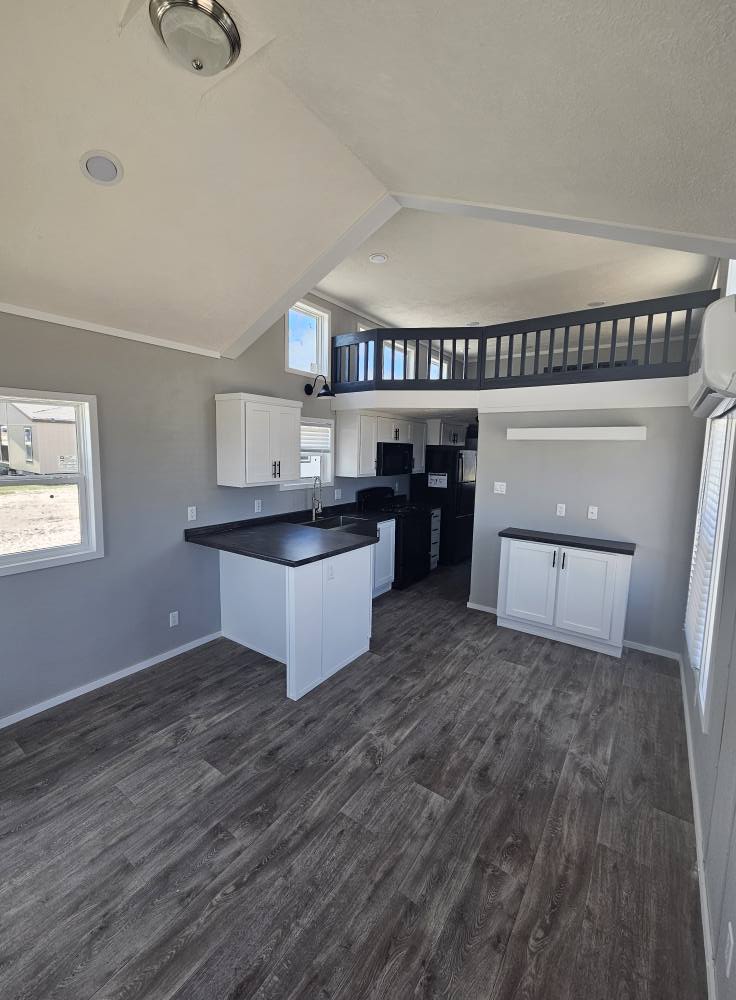 Modern, minimalist tiny house interior with vaulted ceiling, loft, and large windows. It features a compact kitchen, white cabinets, and dark countertops.