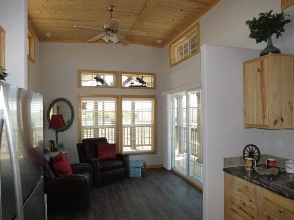 Cozy living room with wooden ceiling and floor. Leather armchairs with red cushions face a window. Natural light fills the space, creating a warm atmosphere.