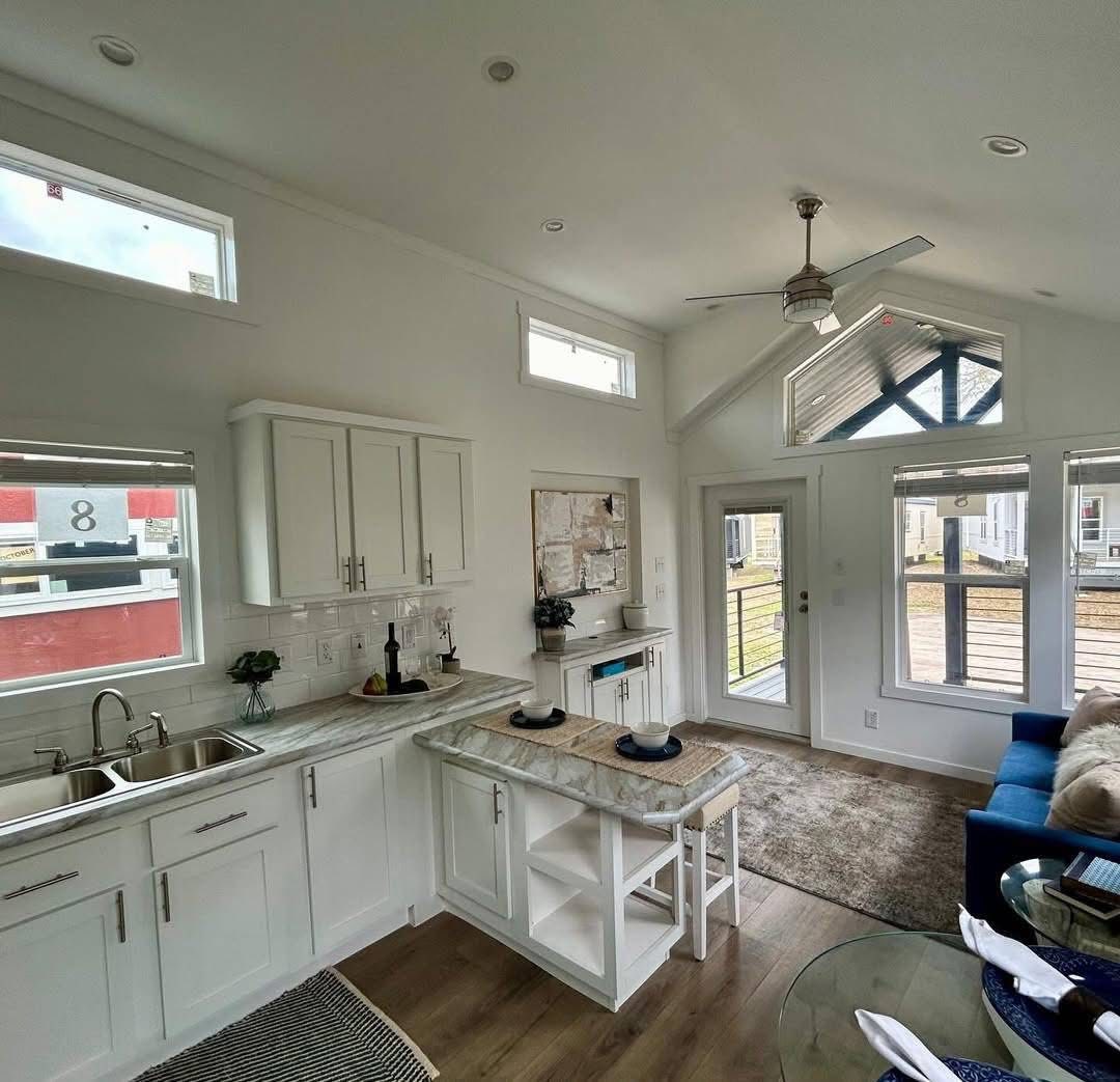 Modern luxury white farmhouse-style park model tiny home interior featuring a bright, open-concept living space with high vaulted ceilings, a contemporary brushed nickel ceiling fan, and energy-efficient transom windows that flood the room with natural light. The sophisticated architectural design showcases white shaker cabinetry, elegant marble-patterned countertops with a built-in breakfast bar and tan upholstered stools, and premium wide-plank wood-style flooring throughout. This professional "full rank" SEO-optimized small-space living solution includes a minimalist built-in media console with marble accents, stylish abstract wall art, and a view into a fully-equipped gourmet kitchen, making it an ideal showcase for custom-built prefab homes, luxury vacation cottages, or modern backyard accessory dwelling units (ADU) designed for maximum style and functional elegance.