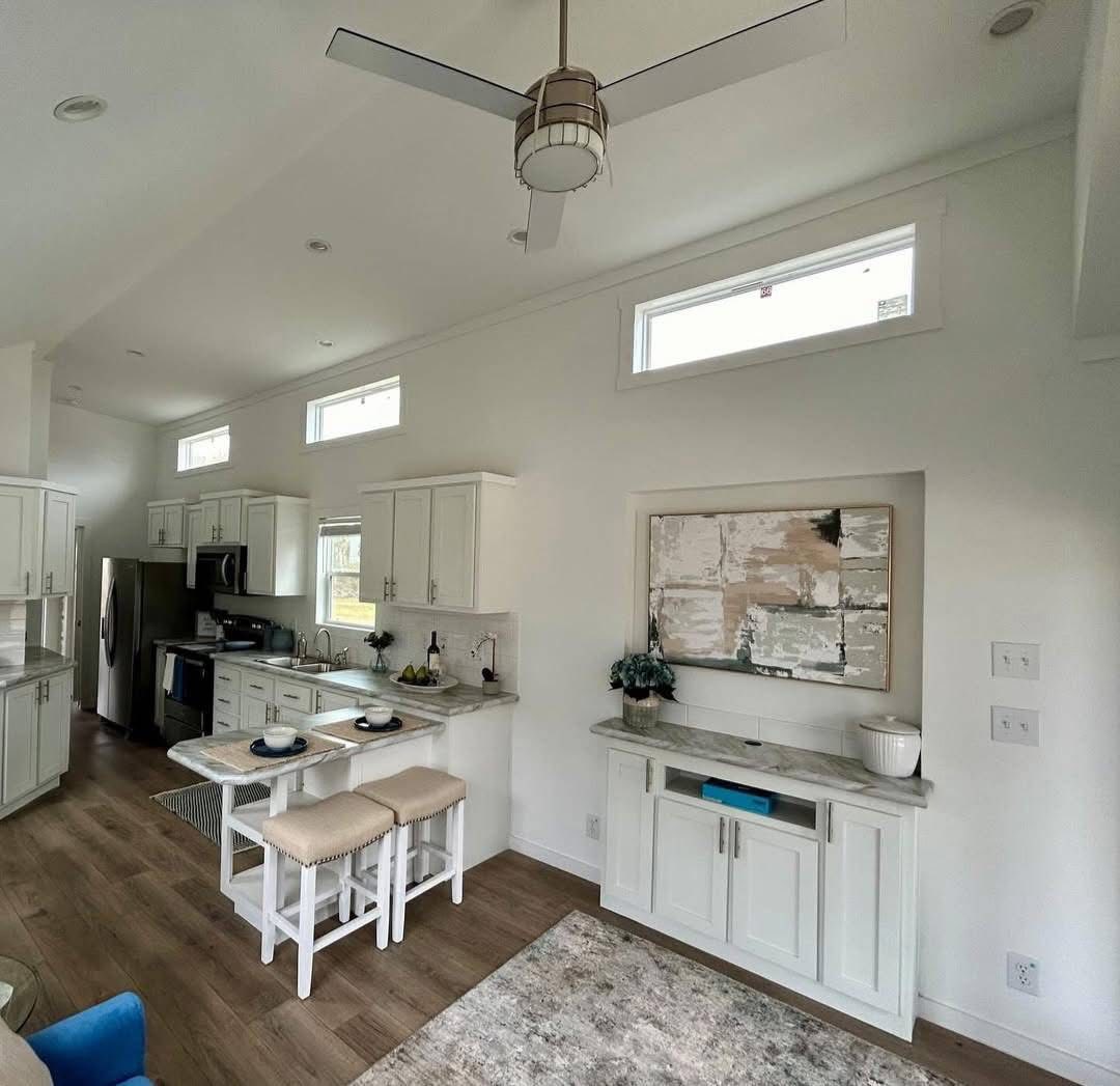 Modern luxury white farmhouse-style park model tiny home interior featuring a bright open-concept kitchen and living area with white shaker cabinetry, marble-patterned countertops, and a cozy breakfast bar with tan upholstered stools. The sophisticated architectural design includes high vaulted ceilings with a contemporary silver ceiling fan, energy-efficient transom windows that flood the space with natural light, and premium wide-plank wood-style flooring. This professional "full rank" SEO-optimized small-space living solution highlights a built-in media console with elegant marble accents and abstract wall art, making it an ideal showcase for custom-built prefab homes, luxury vacation cottages, or modern backyard accessory dwelling units (ADU) designed for maximum style and functional elegance.