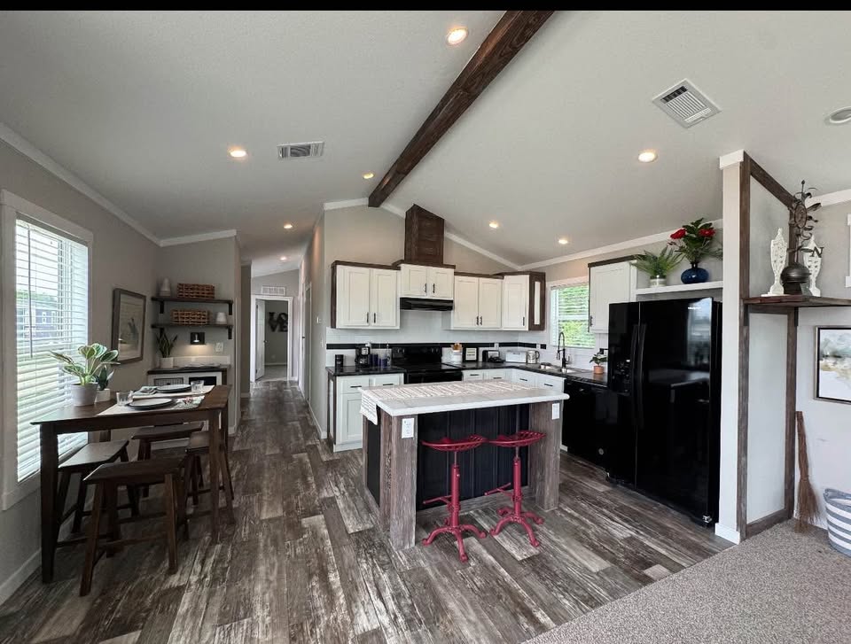 Modern kitchen with rustic wood flooring, featuring white cabinets, a central island with red stools, and a black fridge. Dining area visible to the left.