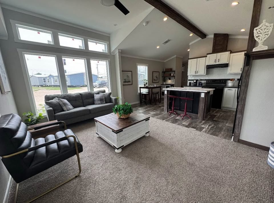 Modern living room and kitchen with large windows, a gray sofa, black armchair, white coffee table, and dark wood accents. Bright and inviting.