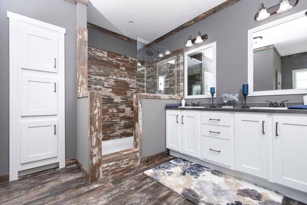 Modern bathroom with rustic wood accents features a glass-enclosed shower, white vanity with dual sinks, large mirrors, gray walls, and decorative lighting.