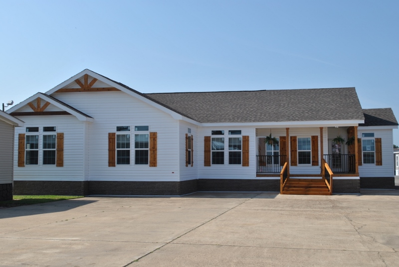 Single-story house with white siding, dark roof, and wooden shutters. Front porch with plants, wooden steps, and a spacious concrete driveway under clear skies.