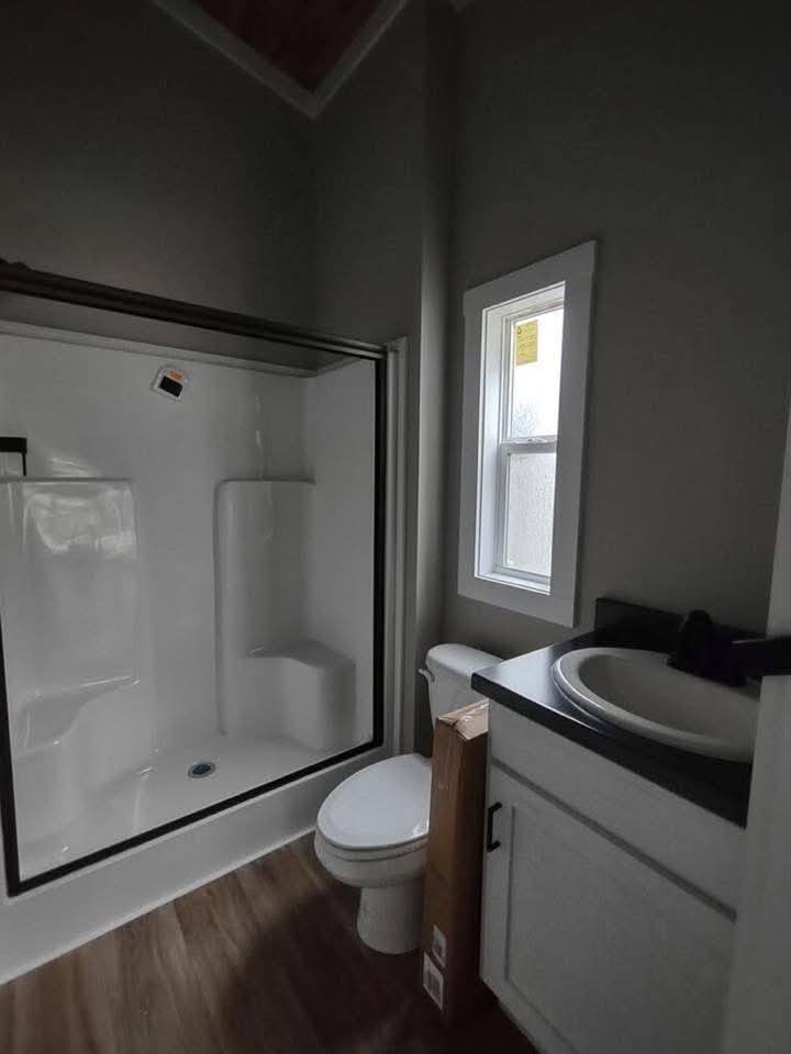 Modern small bathroom interior featuring a clean white walk-in shower with a built-in seat, a white vanity with a sleek black countertop and integrated sink, and a standard white toilet, all set against neutral grey walls with crisp white trim and wood-look vinyl flooring for a functional and contemporary 2-bedroom 1-bath tiny home design.