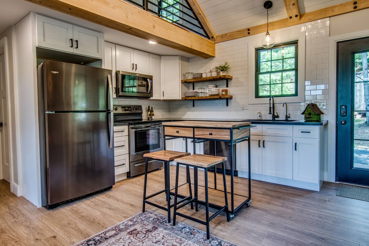 Modern kitchen with white cabinets, stainless steel appliances, and a wooden island with stools. Open shelving and a rustic wood beam add charm.