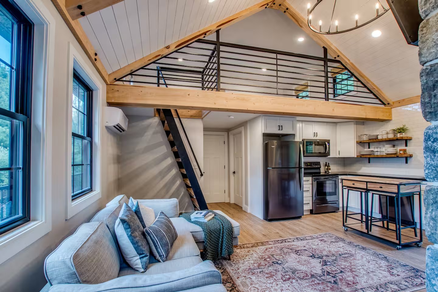 Cozy open-plan living area featuring a loft with railing, modern kitchen, gray couch, wooden accents, and large windows, conveying a rustic yet modern vibe.