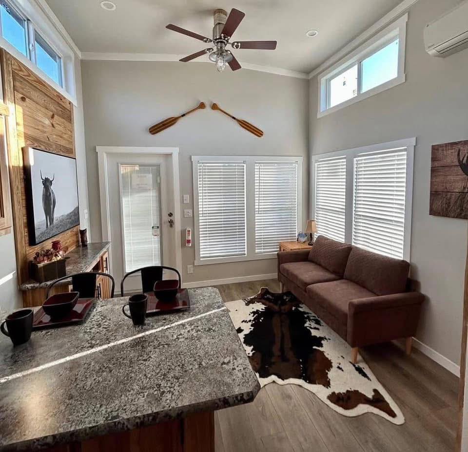 Modern luxury tiny home interior featuring a compact open-concept living area with a brown mid-century modern sofa, a cowhide area rug over high-end wood-look flooring, and a granite-style breakfast bar, accented by rustic wall decor, vaulted ceilings with a classic ceiling fan, and large clerestory windows for a sophisticated move-in ready turn-key residential design.