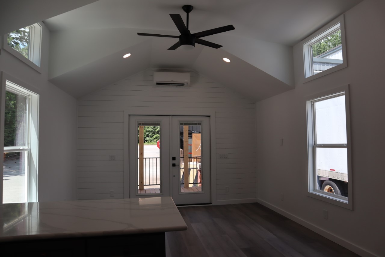 Modern luxury tiny home interior featuring a bright open-concept living area with crisp white shiplap walls and a vaulted ceiling, showcasing a large glass double-door entrance with side windows that flood the space with natural light, complemented by premium grey wood-look flooring, recessed lighting, and a sleek black ceiling fan for a sophisticated and spacious move-in ready turn-key residential design.
