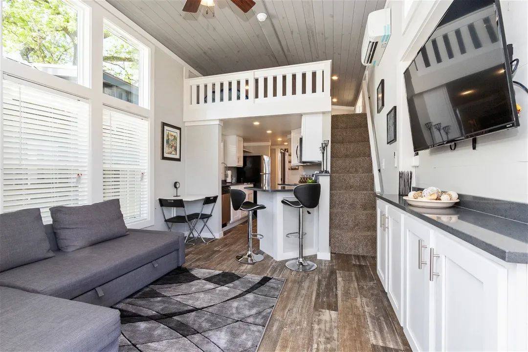 Modern tiny house interior with high ceilings and large windows. Features a gray sectional sofa, kitchen island with stools, loft area, and mounted TV.