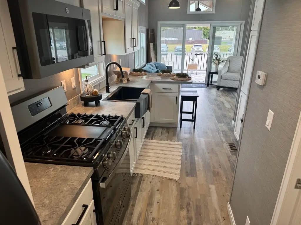 Modern kitchen with light wood flooring and white cabinets. Features a gas stove, black sink, and breakfast bar with stools. Sunlight streams through large windows, creating a warm ambiance.
