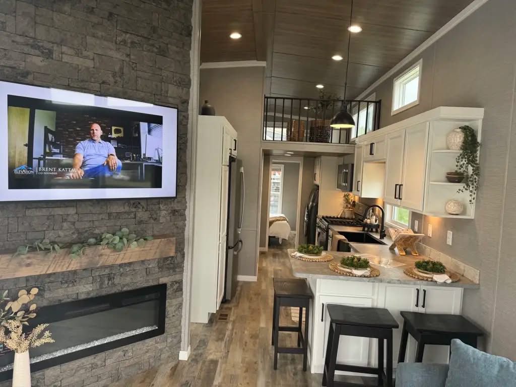 Modern tiny home interior with stone wall and mounted TV. Open kitchen features white cabinets, marble island with stools, and loft. Cozy ambiance.