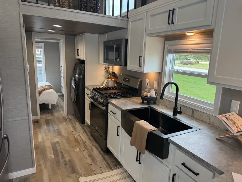 Compact and modern tiny house kitchen with white cabinets, black appliances, and a farmhouse sink. Cozy bedroom visible in the background. Warm, inviting tone.
