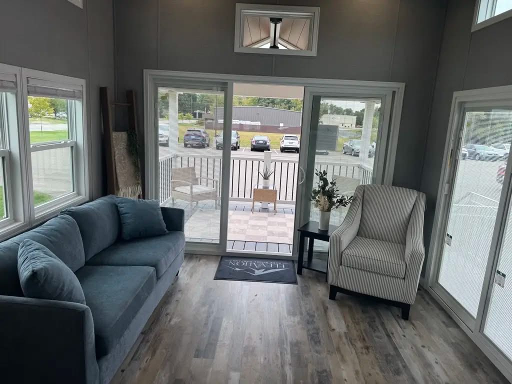 Bright room with large windows, a blue sofa on the left, and a striped armchair on the right. Glass doors lead to a patio overlooking parked cars.