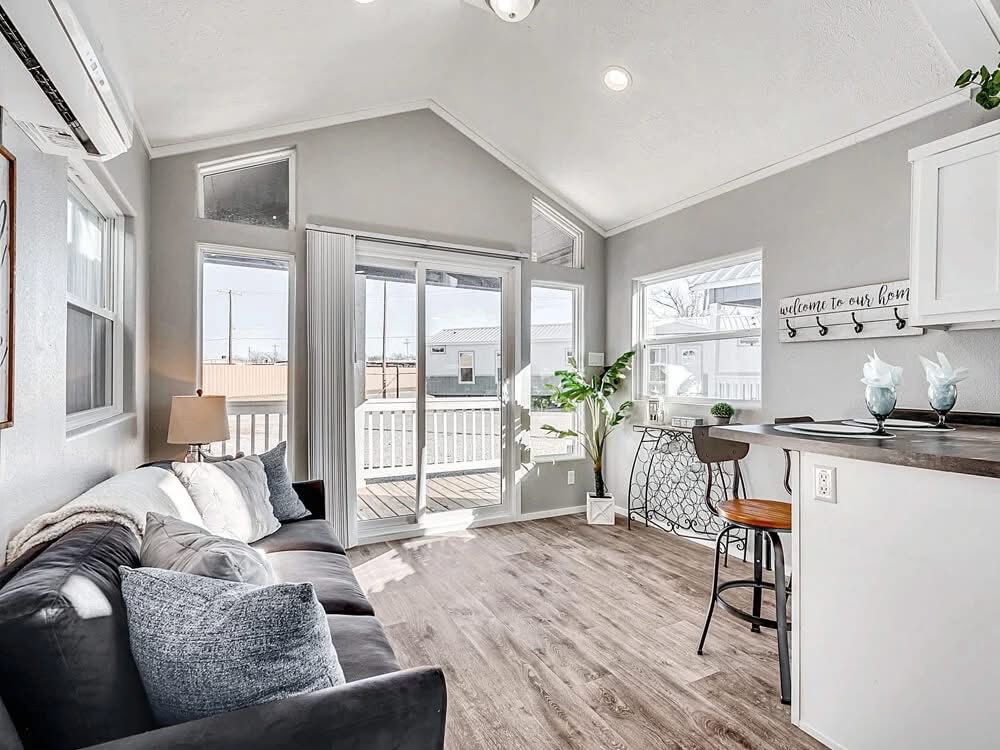 Modern luxury tiny home interior featuring a bright, open-concept living area with high vaulted ceilings, grey walls, and premium wide-plank wood flooring. The sophisticated small-space design showcases a plush dark grey sofa, energy-efficient clerestory windows, and a sleek white kitchen with a breakfast bar and rustic wooden stools. This premium "1 bed / 1 bath" park model captures a professional "full rank" SEO-optimized aesthetic with contemporary lighting, a sliding glass door leading to a private porch, and stylish minimalist decor, offering a perfect high-end prefab living solution for a custom-built vacation retreat or a modern backyard accessory dwelling unit (ADU).