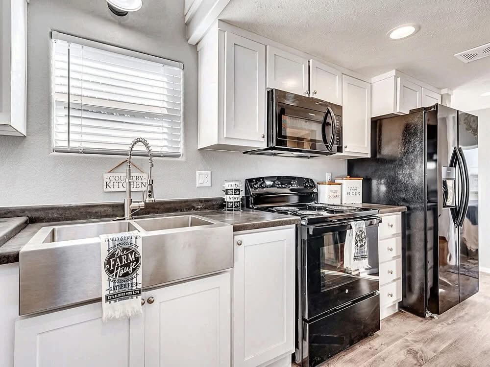 Modern luxury farm-house style tiny home kitchen featuring bright white shaker cabinets, a professional-grade stainless steel apron-front farmhouse sink with a high-arc spring neck faucet, and sleek black appliances including a glass-top range and large side-by-side refrigerator. The sophisticated small-space design is highlighted by dark grey countertops, premium wide-plank wood flooring, and energy-efficient recessed LED lighting, while decorative touches like "Country Kitchen" and "Farm House" signage add a refined rustic charm. This high-end park model interior offers a perfect "full rank" SEO-optimized aesthetic for luxury prefab living, custom tiny house kitchen remodels, or stylish vacation cottage designs with maximum functionality and professional appeal.