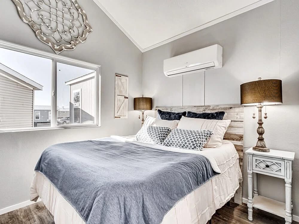 Modern luxury farmhouse-style tiny home bedroom featuring a plush queen-sized bed with a rustic reclaimed wood headboard, layered grey and white bedding, and elegant bronze-finished bedside lamps on a white vintage-style nightstand. The sophisticated small-space interior showcases a high vaulted ceiling with a wall-mounted ductless air conditioning unit, large energy-efficient windows providing abundant natural light, and premium wide-plank wood flooring that enhances the airy, professional aesthetic. This high-end park model design captures a serene "full rank" SEO-optimized atmosphere, making it a perfect example of luxury prefab living, custom-built vacation cottage interiors, or stylish master bedroom solutions for modern accessory dwelling units (ADU).