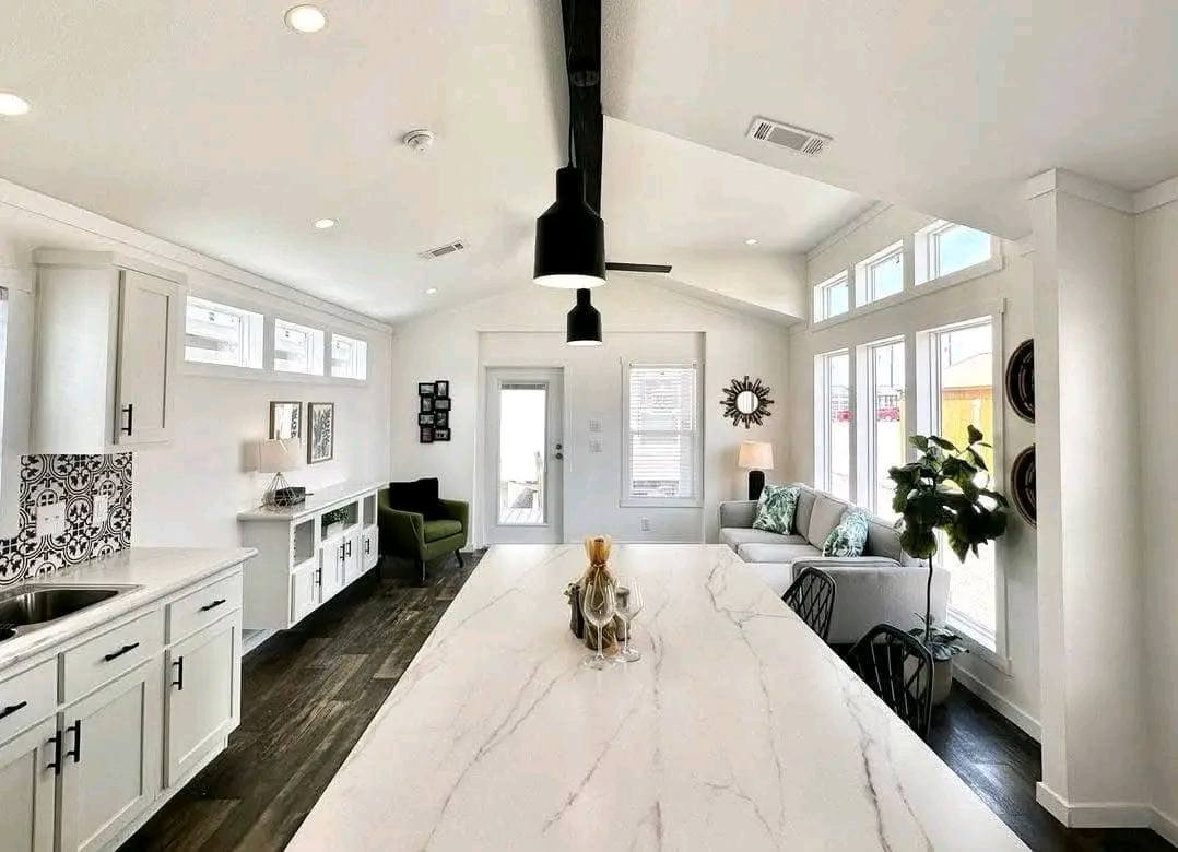 Bright modern kitchen and living area with white walls and cabinets, a large marble island, dark wood flooring, black pendant light, and a cozy sofa.