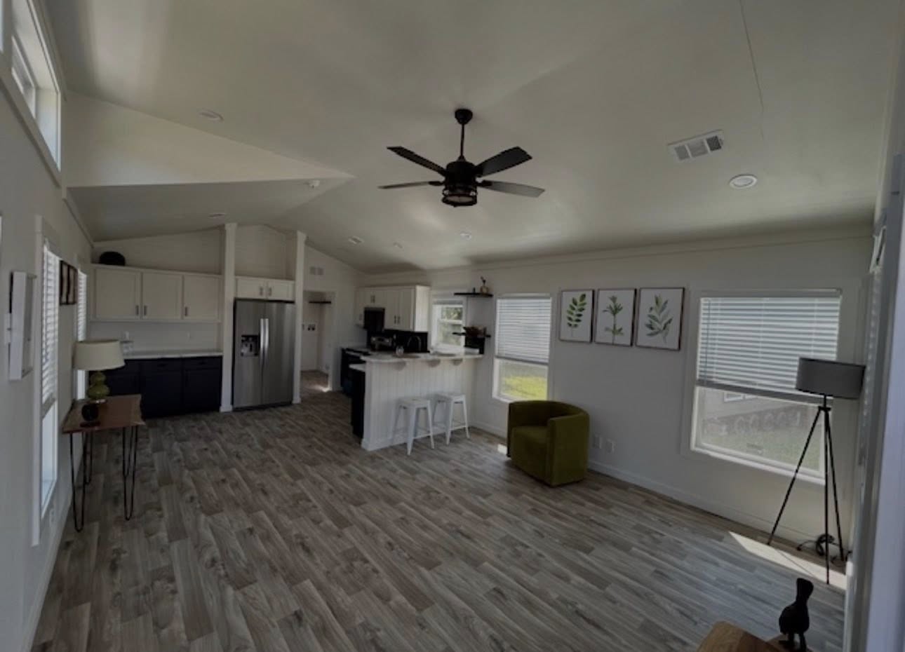 Spacious living room with modern decor. White walls, wood flooring, ceiling fan, green armchair, and framed plant art. Kitchen with stools in background. Peaceful and inviting.
