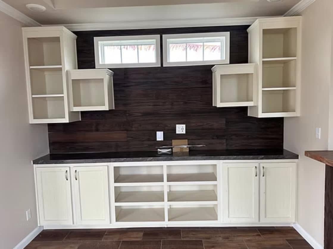 Modern luxury modular home interior featuring a spacious open-concept living area with premium wood-look tile flooring and neutral warm-toned walls, showcasing a high-end white shaker-style cabinetry system with a dark wood-plank accent wall and integrated shelving under a sophisticated tray ceiling with a contemporary fan for a 100% move-in ready turn-key residential design.