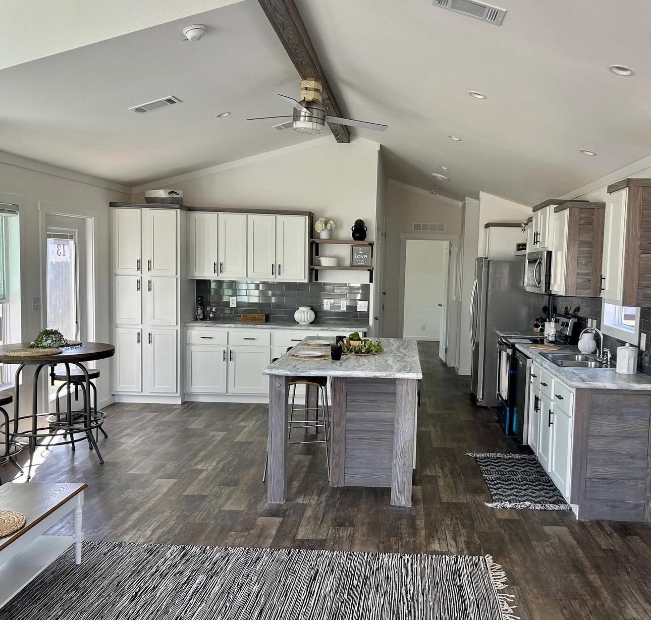 Spacious modern kitchen with vaulted ceilings and exposed beam. Features white cabinets, gray backsplash, island with stools, and stainless appliances.