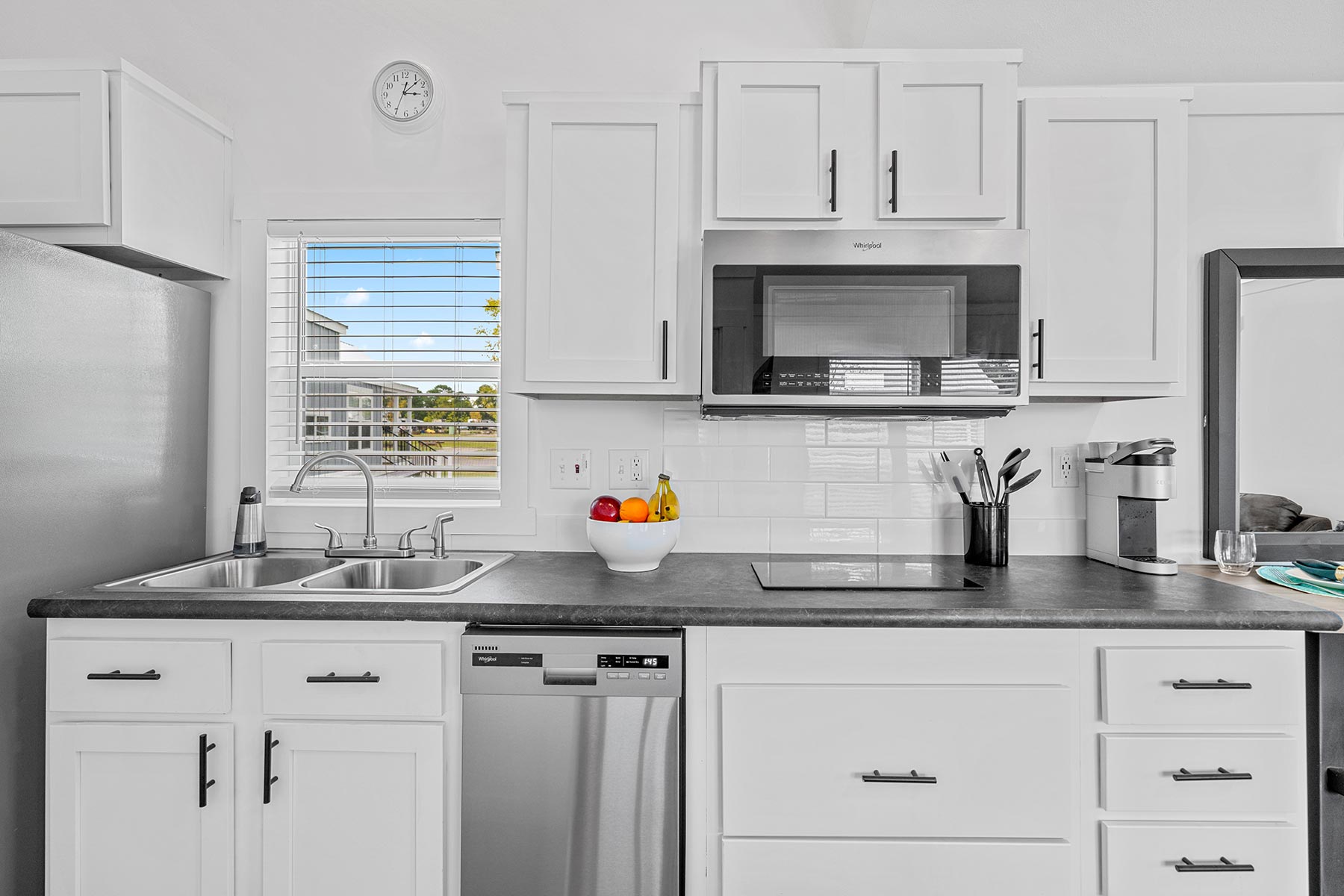 SEO-optimized alt text for a high-ranking luxury park model tiny home kitchen featuring a professional modern interior design with bright white shaker-style cabinetry and a sleek charcoal countertop; the efficient culinary space showcases a premium stainless steel dishwasher, a built-in microwave, and a high-end smooth-top electric cooktop, complemented by a crisp white subway tile backsplash, a double-basin undermount sink with a gooseneck faucet, and a sunlit window with integrated blinds, perfectly highlighting upscale small-space kitchen renovations, luxury prefab tiny house floor plans, and high-end residential mobile living solutions for maximum search engine visibility and ranking.