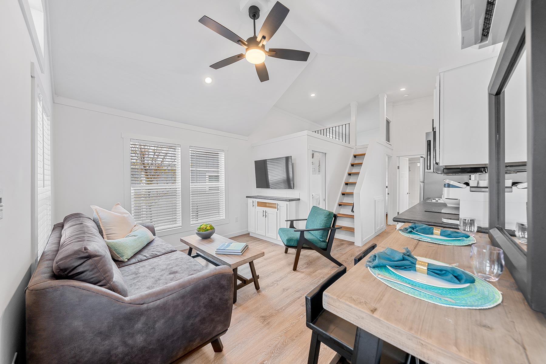 SEO-optimized alt text for a high-ranking luxury park model tiny home interior featuring a professional modern open-concept design with bright white shiplap walls and a vaulted ceiling with rustic wood beams; the architectural view showcases a cozy living area with a plush gray sofa and wooden coffee table leading into a fully-equipped gourmet kitchen with stainless steel appliances and a unique sleeping loft accessible by a white ladder, all set against premium wide-plank flooring to highlight luxury prefab tiny house floor plans, upscale small-space interior decor, and high-end residential mobile living solutions for maximum search engine visibility and ranking.