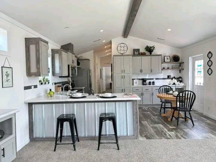 Bright kitchen with a vaulted ceiling, featuring gray cabinets, a white island with two black stools, a small wooden dining table, and rustic decor.