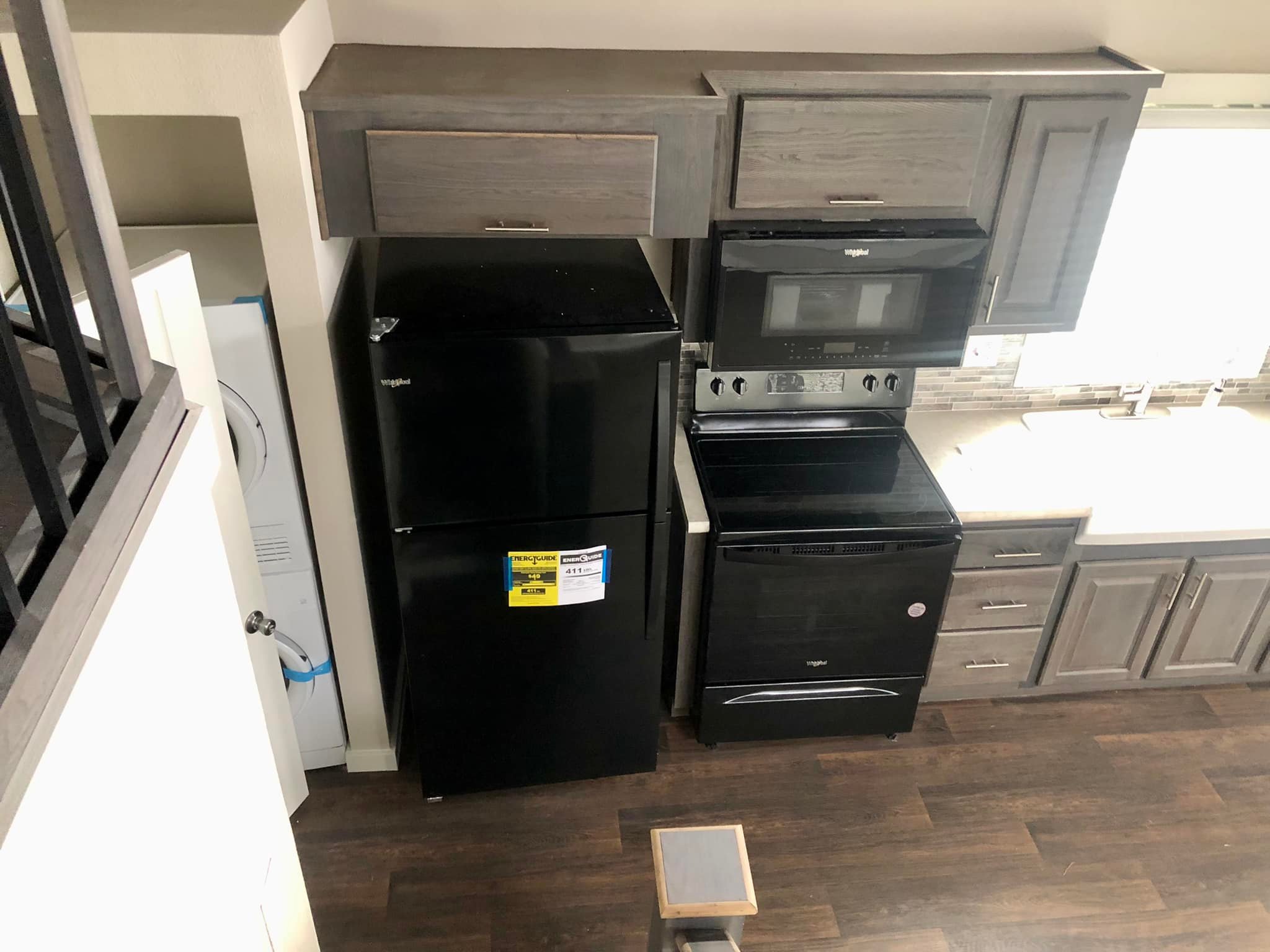 SEO-optimized alt text for a high-ranking luxury custom park model tiny home interior featuring a professional modern kitchen and compact laundry area; the architectural view showcases a high-end black Whirlpool appliance suite including a top-freezer refrigerator, electric range, and over-the-range microwave built into sophisticated gray wood-grain cabinetry, complemented by a space-saving stacked washer and dryer unit, premium dark wide-plank flooring, and a sleek white countertop with an undermount sink to highlight luxury prefab tiny house floor plans, upscale small-space interior finishes, and high-end residential mobile living solutions for maximum search engine visibility and ranking.