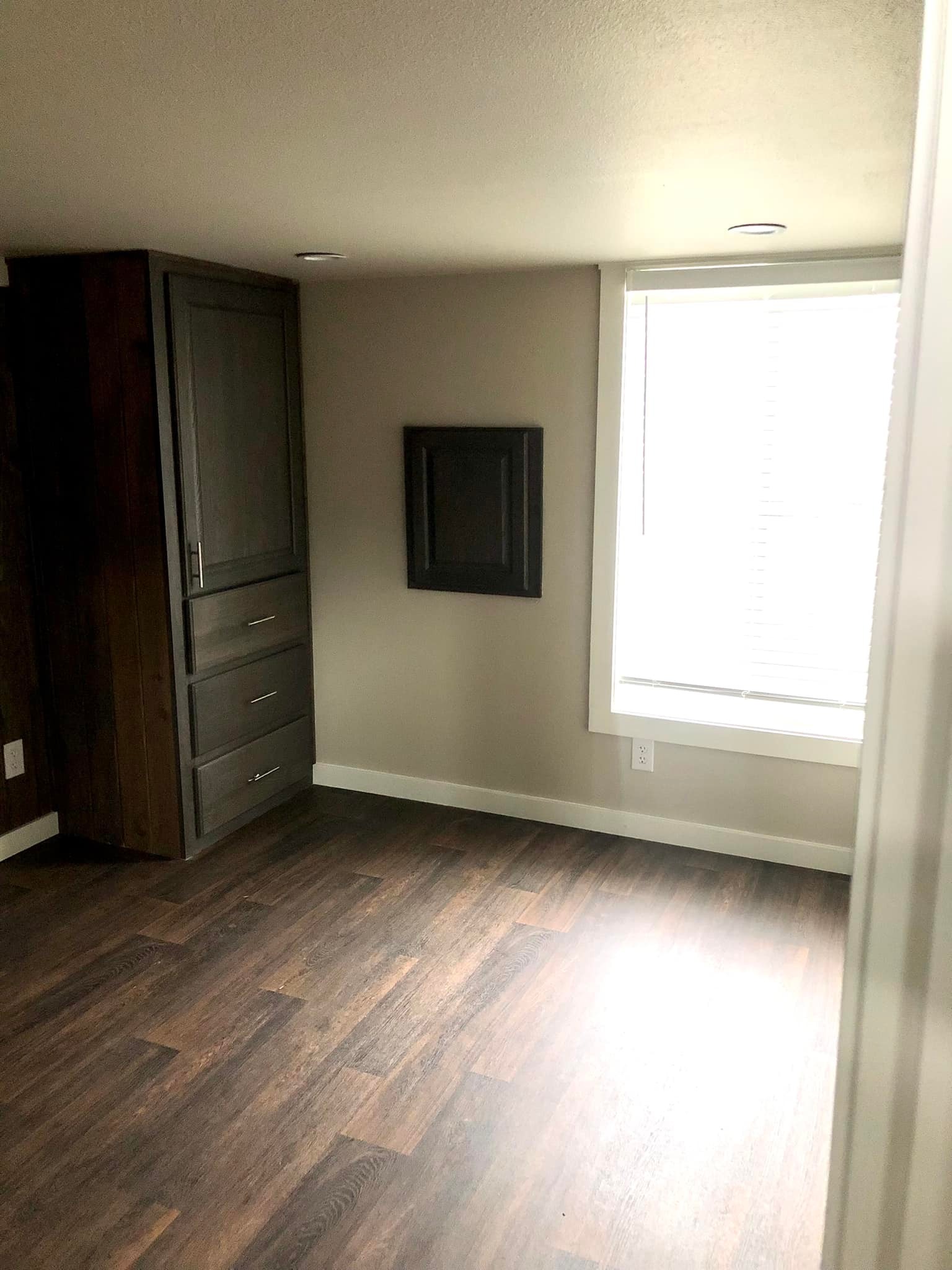 SEO-optimized alt text for a high-ranking luxury custom park model tiny home interior featuring a professional modern master bedroom design with premium dark-wood wide-plank flooring; the space-efficient architectural layout showcases a sophisticated built-in dark-gray dresser and wardrobe unit with brushed nickel hardware, a minimalist wall-mounted flat-screen TV area, and a large sunlit window with integrated blinds that fills the room with natural light to highlight luxury prefab tiny house floor plans, upscale small-space bedroom decor, and high-end residential mobile living solutions for maximum search engine visibility and ranking.