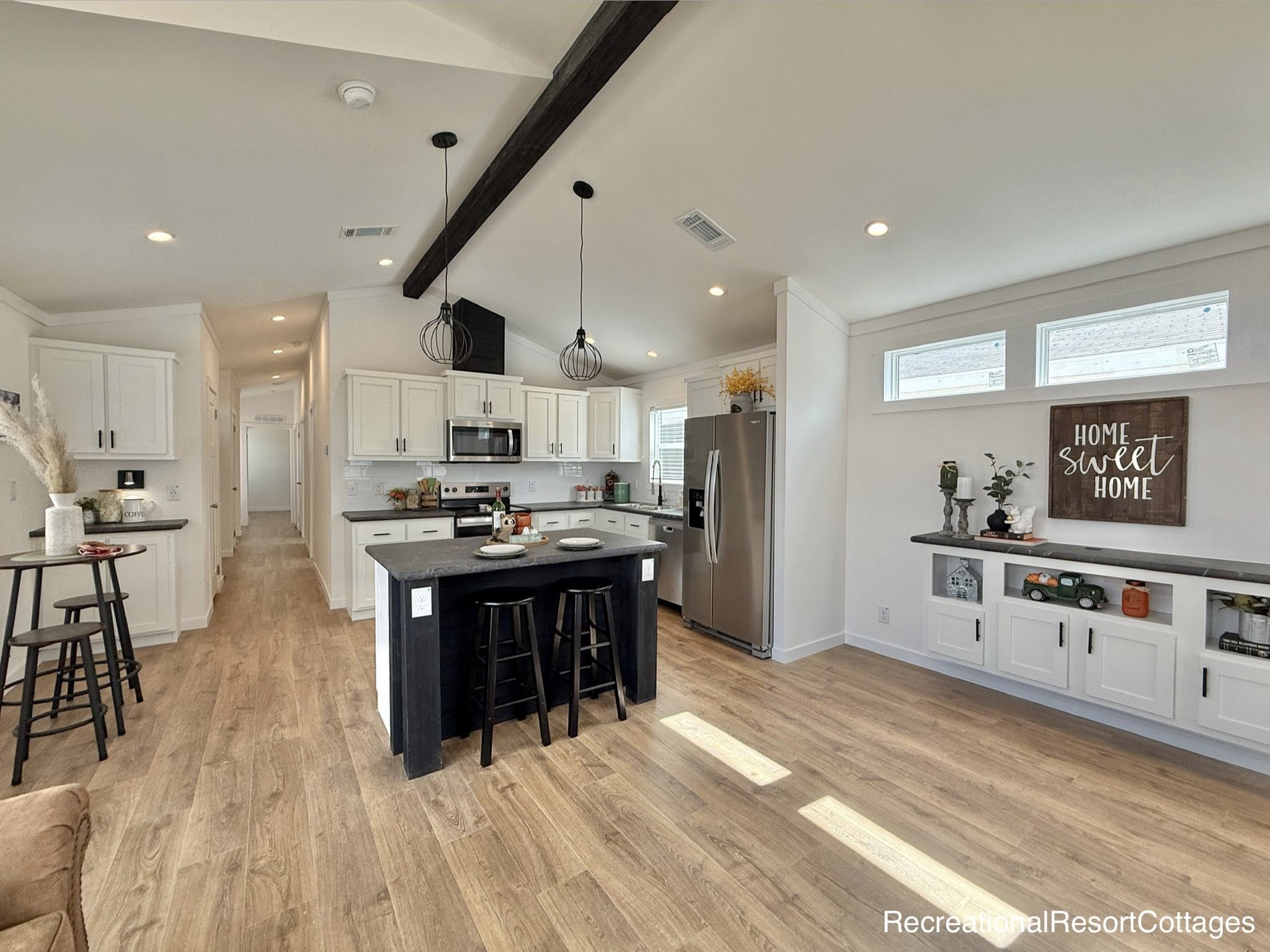Luxury modern farmhouse tiny home interior featuring spacious open-concept layout with vaulted ceiling and dramatic exposed dark wooden beam, recessed LED lighting, black industrial pendant lights over large central kitchen island with dark quartz countertop and black bar stools, white shaker cabinets, stainless steel appliances including side-by-side refrigerator, farmhouse sink, light gray luxury vinyl plank flooring throughout, cozy hallway leading to bedrooms, decorative "Home Sweet Home" wooden sign, built-in shelving with farmhouse accents and decor, bright airy white walls maximizing natural light – premium modular prefab cottage kitchen and living space design ideal for comfortable rural or vacation home living