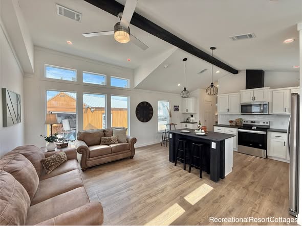 Luxury open-concept tiny home interior with soaring vaulted ceiling featuring exposed black wooden beams, elegant ceiling fan with light fixture, expansive wall of large multi-pane windows flooding space with natural light and scenic outdoor views, light gray luxury vinyl plank flooring, cozy sectional sofa in neutral taupe upholstery with throw pillows, central black island kitchen with quartz countertop, black bar stools, stainless steel appliances, white shaker cabinets, modern pendant lights, recessed ceiling lights, farmhouse-style living room and kitchen combo perfect for modern tiny house, modular home, or prefab cottage living