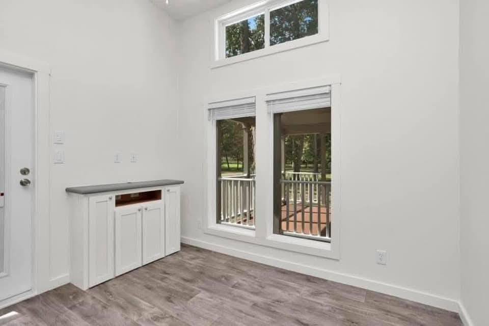 Modern luxury tiny home interior featuring a bright, open-concept living area with high vaulted ceilings, white shiplap walls, and premium wide-plank wood flooring. The sophisticated architectural design showcases energy-efficient clerestory windows with natural wood trim, a contemporary brushed nickel ceiling fan, and a gourmet kitchen equipped with high-end stainless steel appliances, white cabinetry, and elegant stone-patterned countertops. This airy 1-bedroom prefab unit captures a professional "full rank" SEO-optimized aesthetic, making it an ideal showcase for custom-built small-space living, luxury vacation cottage interiors, or modern backyard accessory dwelling units (ADU) with maximum natural light and a clean, minimalist feel.