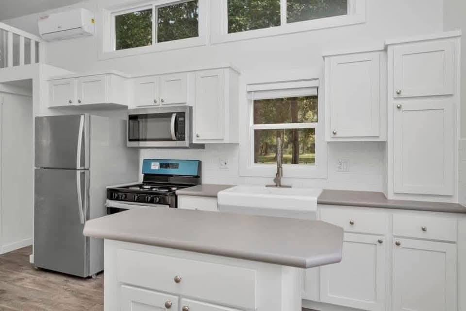 Modern luxury farmhouse-style tiny home kitchen featuring bright white shaker cabinetry, a portable center island with a minimalist grey countertop, and high-end stainless steel appliances including a large refrigerator, gas range, and integrated microwave. The sophisticated architectural design showcases a high vaulted ceiling, energy-efficient clerestory windows, and a spacious loft area accessible by a built-in staircase, all complemented by premium wide-plank wood flooring and clean white shiplap walls. This professional "full rank" SEO-optimized interior offers an ideal inspiration for luxury prefab living, custom-built park model tiny houses, or stylish small-space kitchen designs with maximum natural light and a contemporary, airy aesthetic.