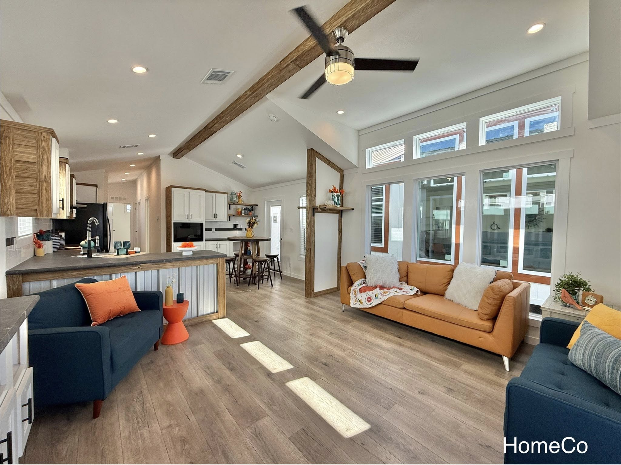Bright, modern open-concept living space with wood beams, ceiling fan, and large windows. Features blue and tan sofas, a kitchen island, and cozy decor.