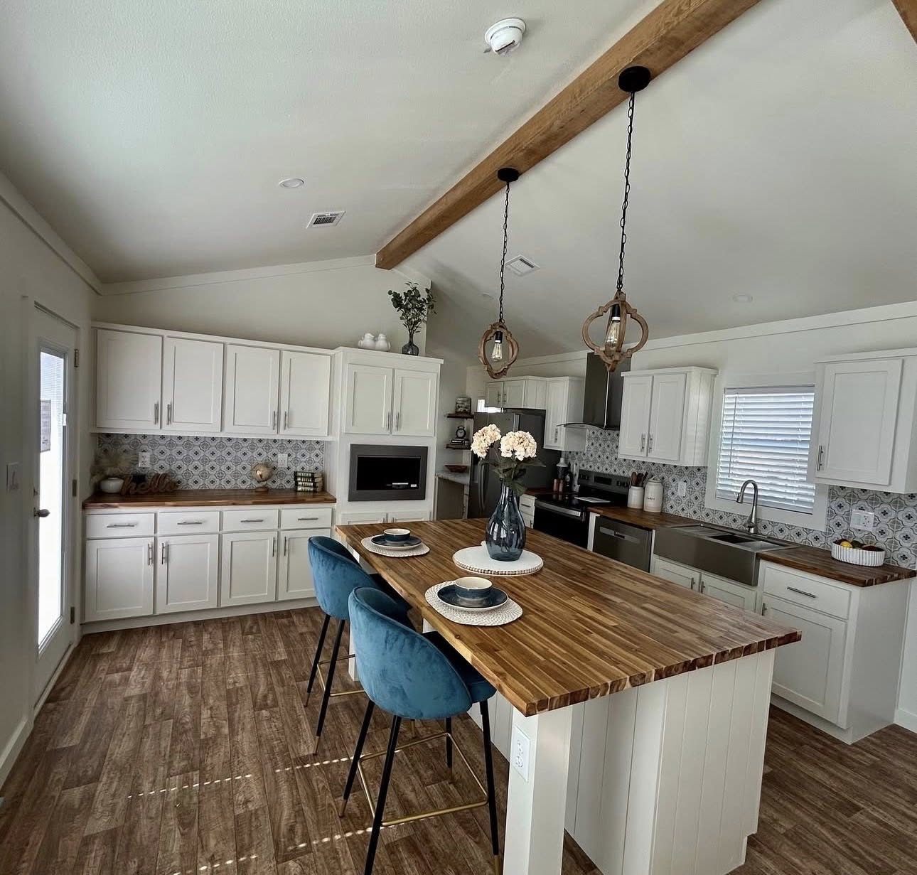 Bright kitchen with white cabinets, a wood island, and blue velvet stools. Pendant lights hang above, and a vase of flowers adds elegance. The space feels modern and inviting.