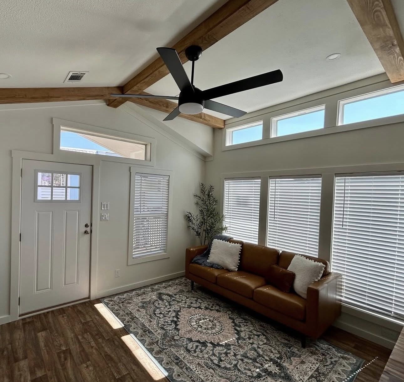 A bright living room with wood ceiling beams and a modern fan. A brown leather sofa with pillows sits on a patterned rug, surrounded by tall windows.