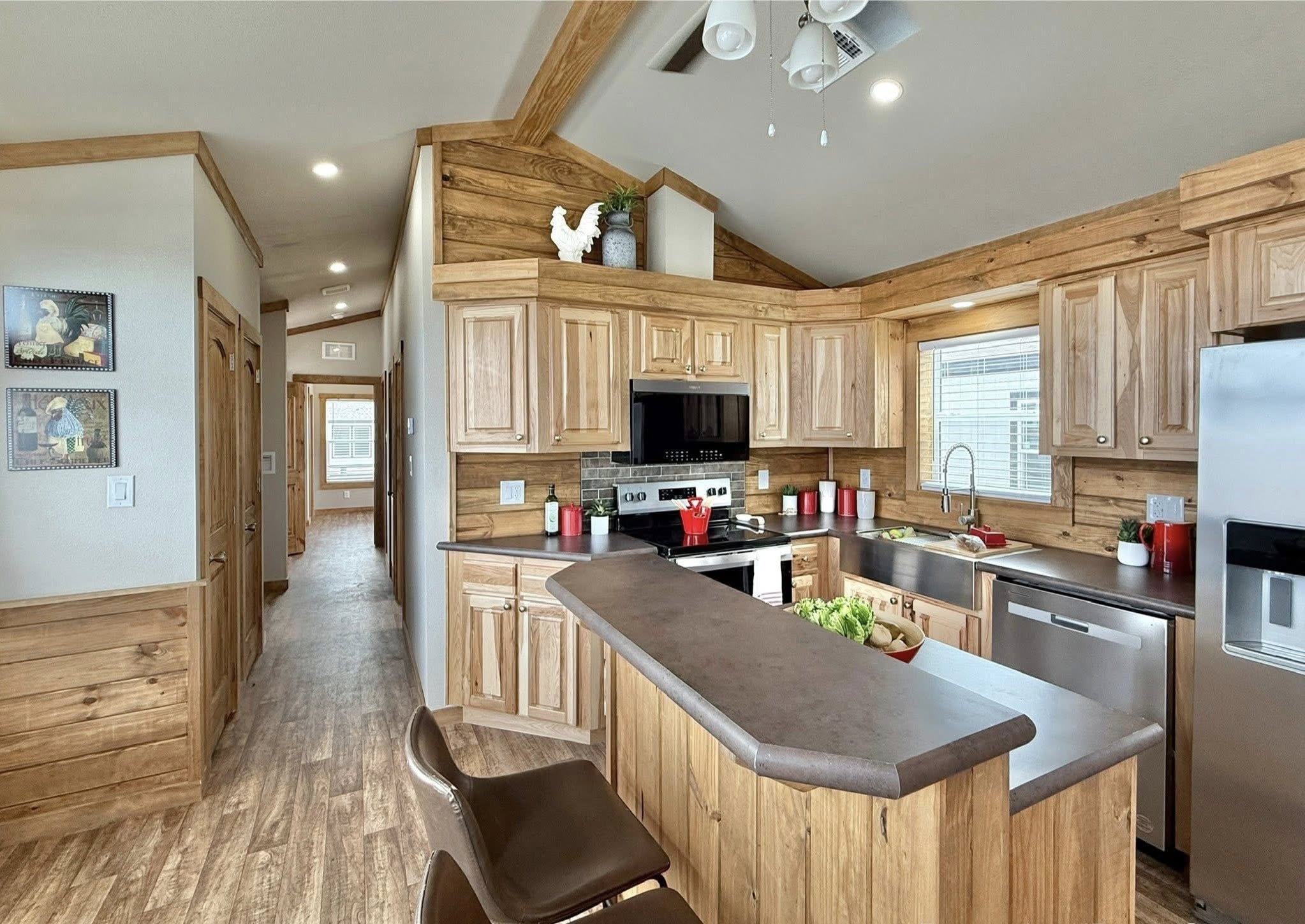 Modern luxury tiny home interior featuring a spacious open-concept kitchen and dining area with high-end natural wood cabinetry, stainless steel appliances including a farmhouse sink, and a sleek grey breakfast bar, all set under vaulted wood-plank ceilings with rustic beam accents and premium wood-look flooring for a sophisticated move-in ready turn-key residential design.