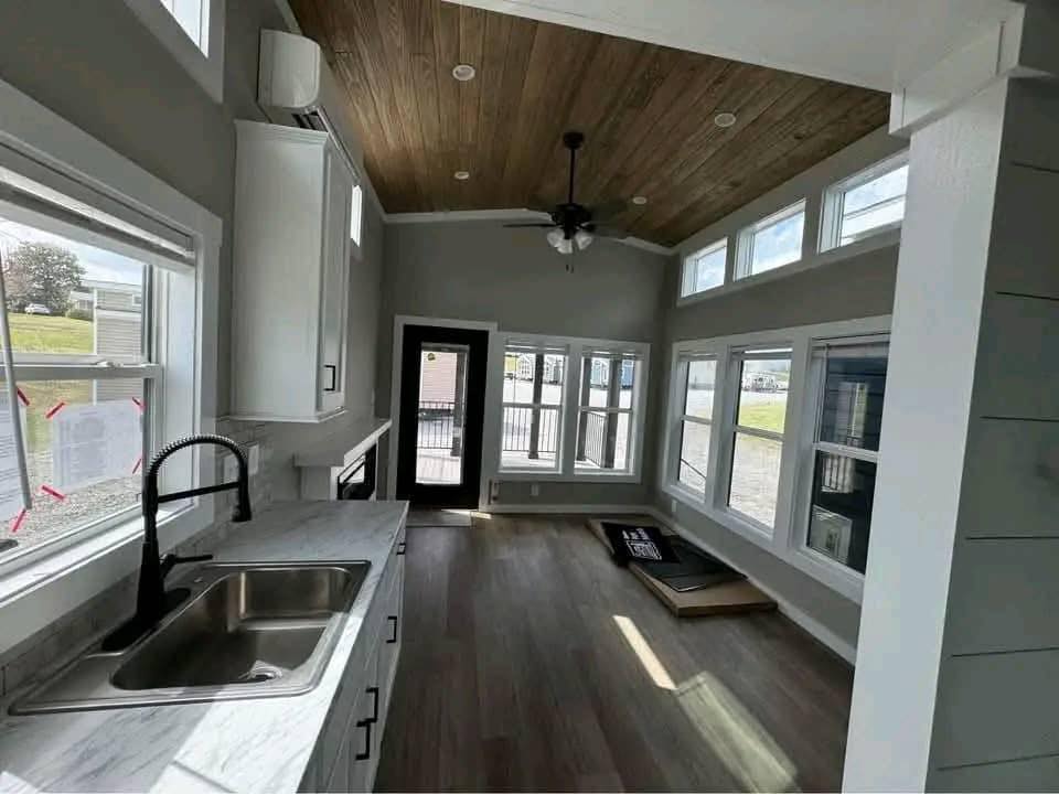 Spacious bright modern farmhouse tiny home interior featuring elongated open-concept kitchen and living layout with vaulted ceiling accented by warm wooden plank paneling, black ceiling fan with lights, recessed LED lighting, light gray shiplap walls, luxury vinyl plank flooring in rich wood tones, white shaker cabinets with gray quartz countertops, large farmhouse sink with matte black gooseneck faucet, abundant natural light from floor-to-ceiling windows and clerestory transoms overlooking scenic outdoor views, black French doors leading to porch, airy minimalist design perfect for cozy tiny house living, off-grid retreats, or compact family-friendly modular homes.